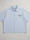 A short-sleeve button-up shirt with vertical light blue and white stripes, featuring a small embroidered logo and text on the left chest, laid flat against a textured light gray background.